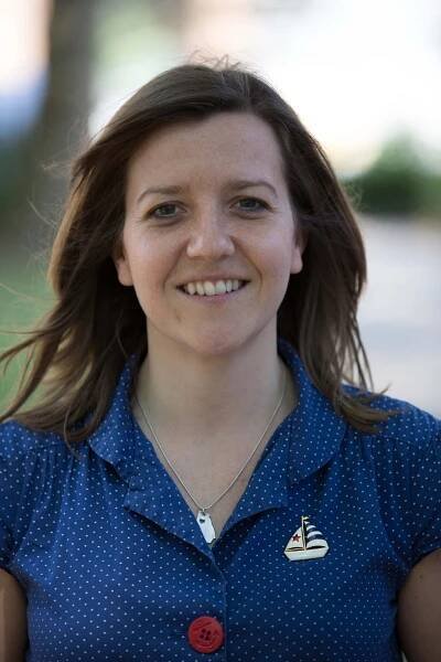 Laura Harrison - White woman with long brown hair wearing a mid blue shirt with a sailing boat motif and red button.