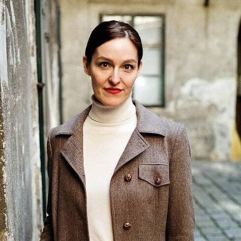 Frau mit rotem Lippenstift und weißer Rollkragenbluse unter einem braunen Mantel an einer Mauer.