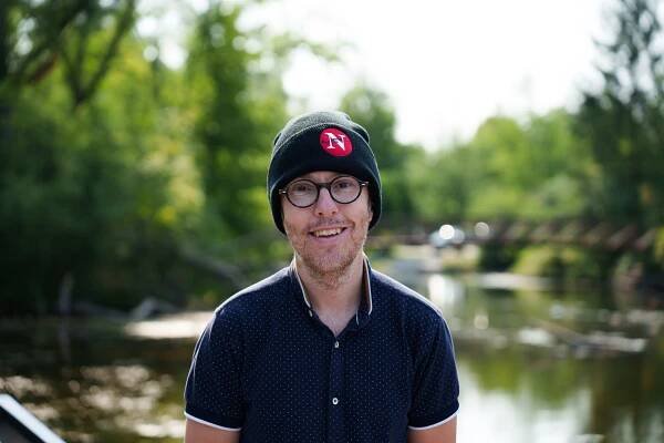 Will Pearson photographed wearing a black Narwhal toque with red logo and a navy t-shirt, against a green natural backdrop. 
