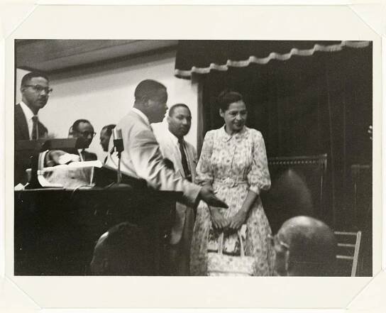 A black-and-white photograph of Ralph Abernathy speaking with Rosa Parks while Martin Luther King Jr. stands in the background. There are several onlookers near a podium to the right of Abernathy and some individuals sitting in chairs in front of the podium. The print is signed [Charles Moore] on the back in pencil.