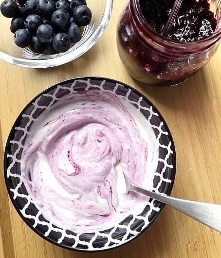 A glass bowl with fresh blueberries next to a jar of blueberry jam, next to a black and white bowl containing blueberry jam swirled into white yogurt.