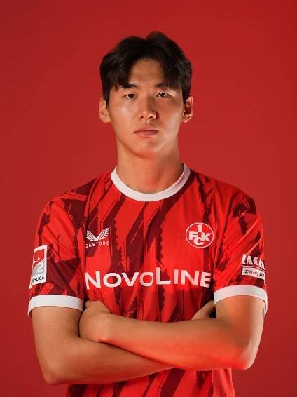 Ein junger asiatischer Mann steht vor einem roten Hintergrund. Er trägt ein rotes Fußballtrikot mit verschiedenen Sponsorenlogos und blickt ernst in die Kamera.