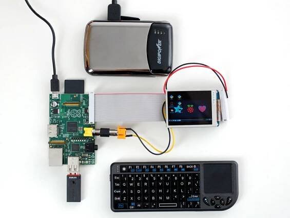 A silver rectangular USB battery pack powering a mini wireless keyboard, a green microcontroller, and a full-size breadboard with graphic display breakout.