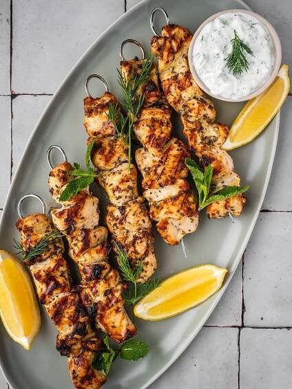 Greek chicken souvlaki skewers on a platter garnished with dill, mint, sliced lemons and a side of yogurt sauce.