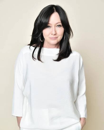 Shannen Doherty poses for a portrait. She has her hair down and is wearing a white blouse with matching white slacks. 