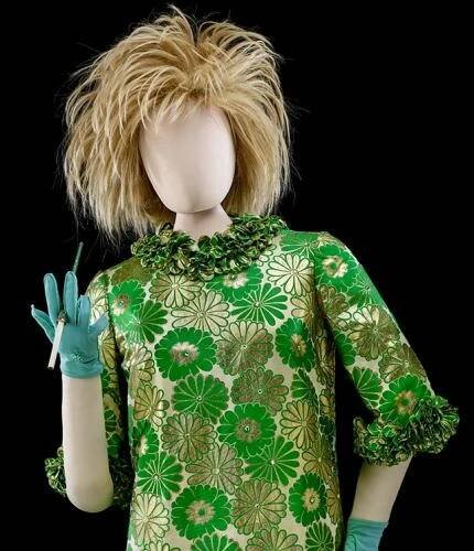 A mannequin wears a blonde, spiky wig and a lacquered shift dress with green and gold daisies. The bottom hem, neckline, and sleeves are ruffled with teal, wrist-length gloves and a cigarette holder. Her shoes are green ankle-high booties with low heels and gold details.