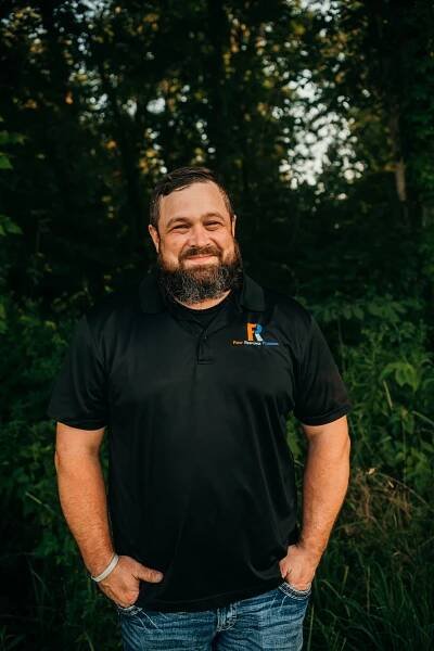 A man with a beard is standing in front of a forest with his hands in his pockets.