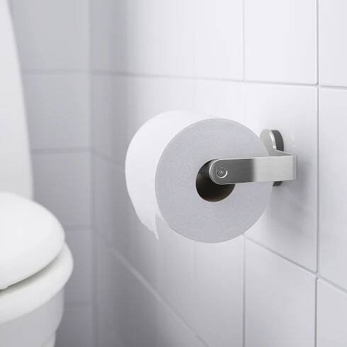 Bílý držák toaletního papíru namontovaný na bílé kachlové stěně, držící roli toaletního papíru, součást série BROGRUND se skrytými šrouby.