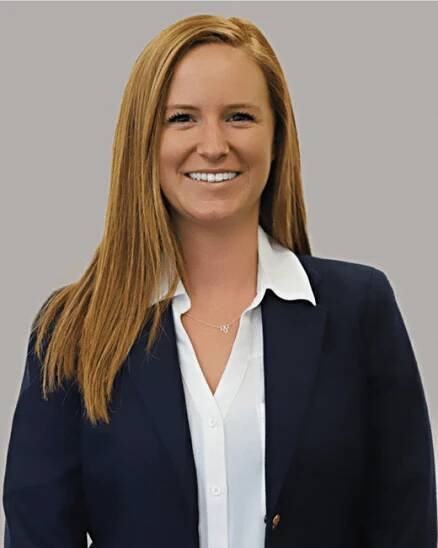 Person with long red hair wearing a navy blazer and white blouse, smiling at the camera against a gray background.