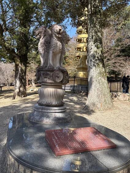 A stone pedestal featuring a sculpture of two lion-like figures, surrounded by trees in a serene park setting. In the background, a golden pagoda is visible, adding to the cultural ambiance.
