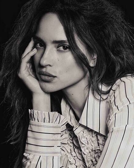 Adria Arjona leaning on her hand in a striped and ruffled shirt.