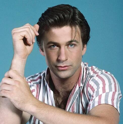 Person sits casually, wearing a striped shirt with rolled sleeves and jeans, posing thoughtfully against a plain backdrop