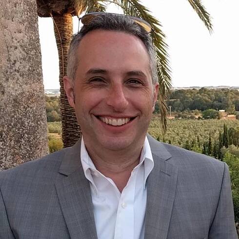 Headshot photograph of Scott Schwartz, who is wearing a white collared shirt under a grey blazer jacket. He is smiling and standing underneath a palm tree.