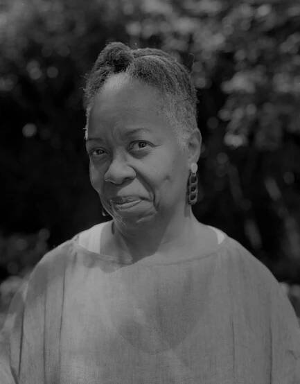 A black-and-white, head-and-shoulders portrait of a woman whose hair, graying at the sides, is pulled up. She is smiling with her mouth closed, revealing dimples. She has a dangly earring.