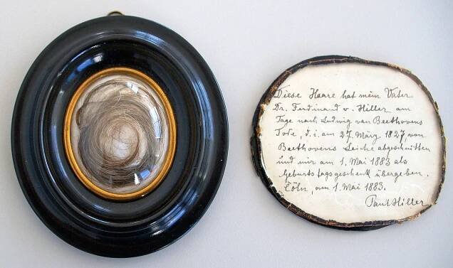 A close-up view of a swirl of reddish-gray hair sits in a locket, left, next to a small circular piece of paper with frayed edges, bearing cursive handwriting in German, signed “Paul Hiller.”