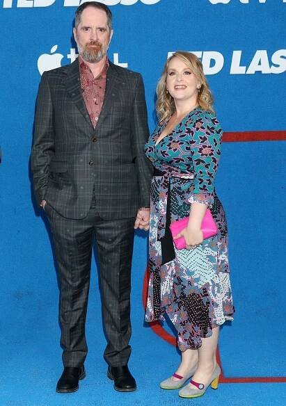 Ted Lasso co-creator Brendan Hunt keeps it quirky in a checkered suit with floral embroidery as he poses with wife Shannon Nelson, who adds a playful pop of color in a floral wrap dress, colorblock heels with yellow soles, and a hot pink clutch