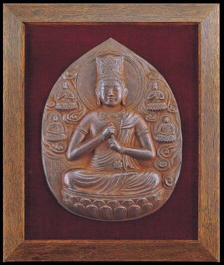 A tear-shaped metallic relief of a buddha wearing a tall crown sitting cross-legged, grasping his upturned left index finger with his right hand, surrounded by clouds and four smaller buddhas.