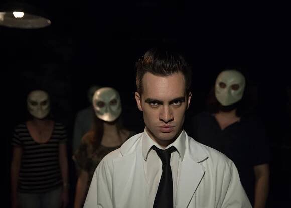 Brendon Urie stairs straight ahead in "Sleep No More."  Masked audience members stand behind him. 