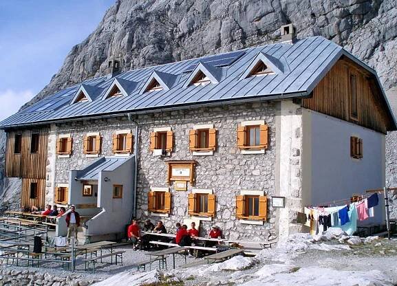 Berghütte mit Steinmauern und einem Metalldach, umgeben von felsigem Gelände. Die Menschen sitzen an Tischen im Freien, in der Nähe hängt Wäsche.