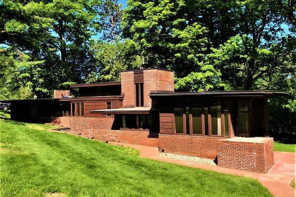 An exterior view of a brick Usonian Frank Lloyd Wright house with green grass in the front and trees in the back.