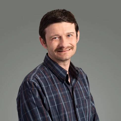 Daniel Miller, CPS, MPA, Business Manager at the Institute@MHP in Philadelphia, smiling in a black sweater and plaid shirt against a light gray background, representing Mental Health Partnerships’ commitment to peer-led mental health training.
