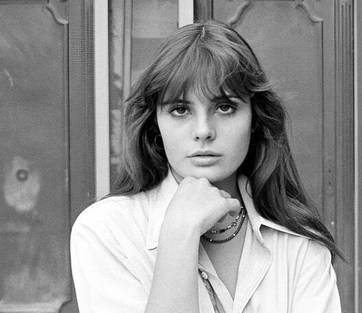 Marie Trintignant pózuje při pohledu do fotoaparátu bez zvláštních emocí. Nosí dlouhé hnědé vlasy a ofinu. Pravou rukou se dotýká krku, paži složenou před sebou. Je oblečená v bílé halence a na krku má náhrdelník z ozdobných dřevěných korálků. Foto: Etienne George / Volné dílo
