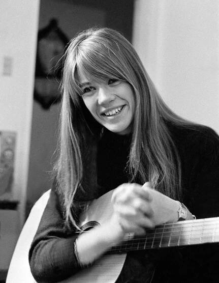 Françoise Hardy holds a guitar and smiles, her long hair falling over her shoulders.