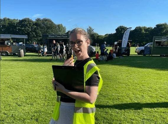 Gina Jackson holding a clipboard and talking into a walkie talkie. The sky is a bright blue behind her and the grass is bright green. The food and drink vans are behind her. 