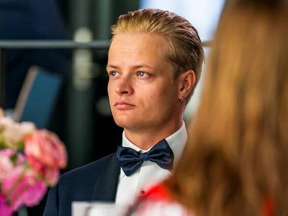 A young man with blond hair, fair skin and blue eyes sitting at a table in a dinner suit.