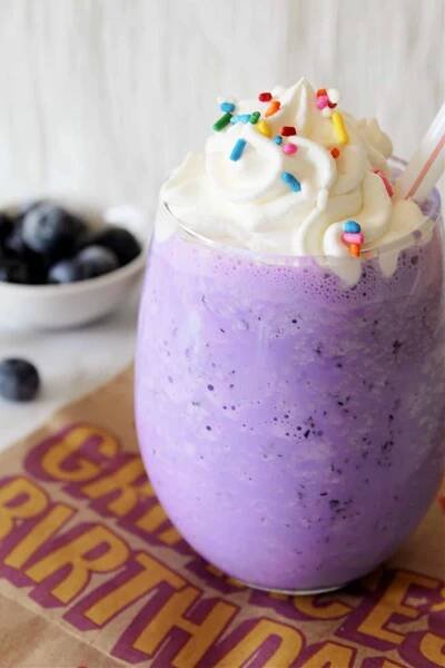 Headon photo of Copycat McDonald's Grimace's Birthday Milkshake with whipped cream and sprinkles. The shake is on top of a McDonald's Grimace Birthday bag. There is a bowl of blueberries in the background.