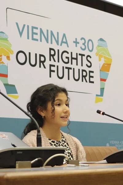 Child human rights defender Dulce Maria from Ecuador is speaking in front of a microphone at a panel during the 2023 Vienna Conference on Youth and Child Human Rights Defenders. Behind her is a screen reading "Vienna +30, Our rights, our future".