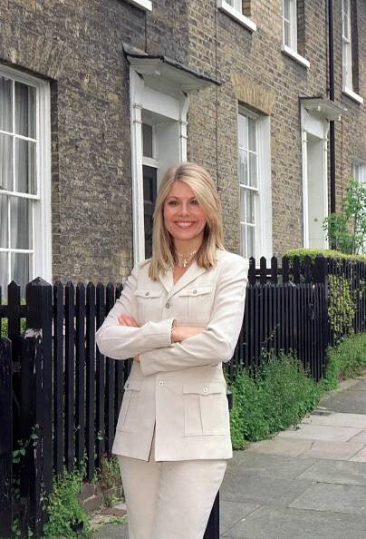 Glynis Barber in a beige suit, standing outside a row of houses.