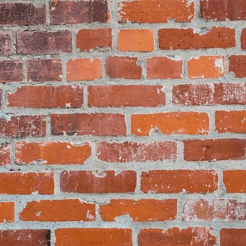 A close-up of a brick wall with deteriorating mortar joints. The mortar appears worn and eroded, with visible cracks and gaps between the bricks. Some bricks show signs of chipping and surface damage. This type of deterioration is often caused by winter weather, as freeze-thaw cycles weaken the mortar, leading to structural issues and increased water infiltration into the masonry wall.