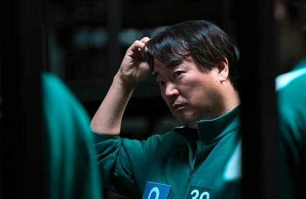 Person in green sports jacket with number, looking serious, hand on head. The setting appears tense, possibly from a popular series