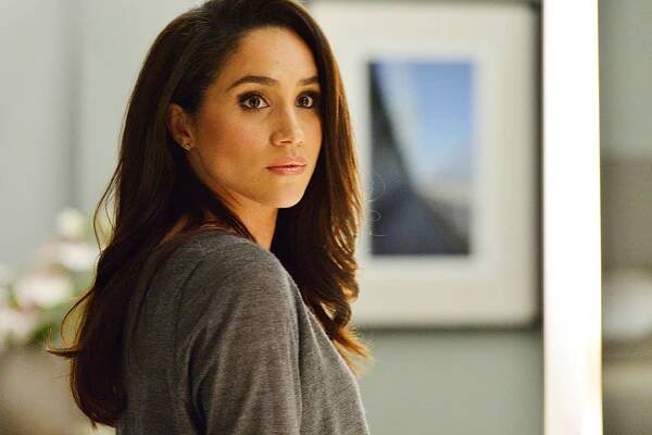 Meghan Markle in Suits with long hair wearing a simple, elegant top, gazing thoughtfully over their shoulder in a softly lit room with framed artwork