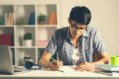 A young man in glasses sits at a table writing in a notebook. For College Application Essays.