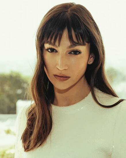 a person with long brown hair wearing a white top set against a blurred outdoor background