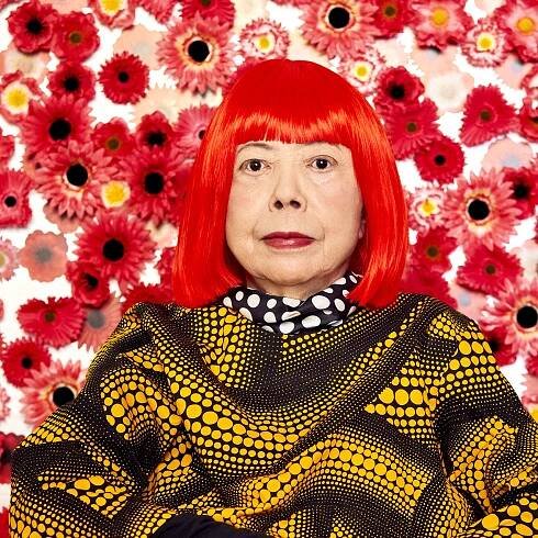 Yayoi Kusama, sporting bright red hair and a black shirt covered in light orange polka dots, sits in front of a white wall covered in red and white flowers.