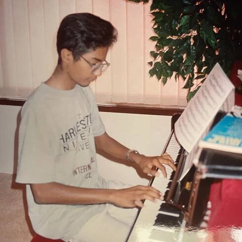 A teenage Jeremy Fernandez plays a small upright piano in a family home. He wears glasses and a tshirt.