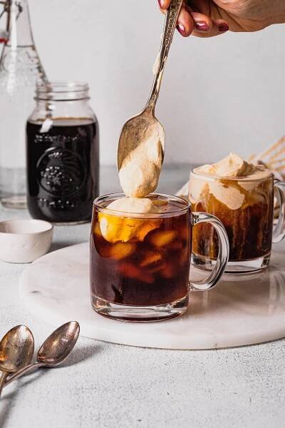 A hand holds an antique silver spoon filled with pumpkin cream cold foam and tops a glass of cold brew coffee with it.