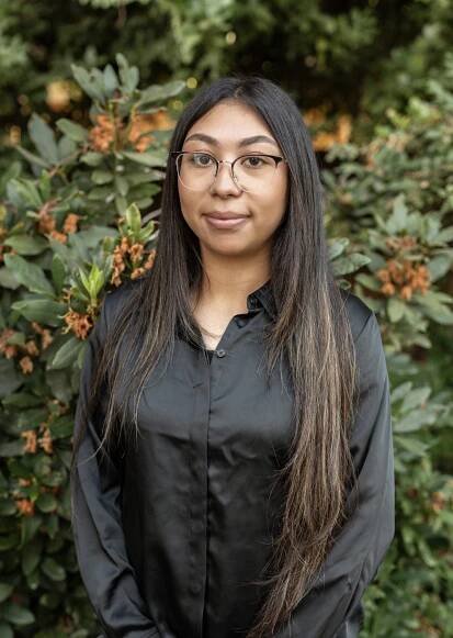 A person with long, dark hair and glasses stands in front of leafy greenery, wearing a black shirt. They have the composed look of someone who might be a dentist in Yuba City, gazing at the camera with a neutral expression.