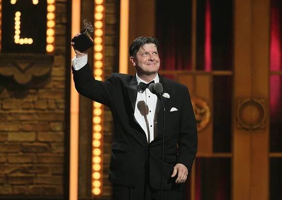 Michael McGrath, wearing a tuxedo and grinning, his hair tousled, stands on a stage and holds up a Tony Award.