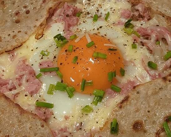 A cracked egg on a French galette bretonne pancake, topped with cheese, ham and chives.