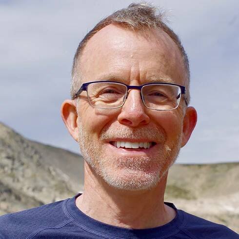 Sunlit head-and-shoulders of a smiling man with short hair and glasses, standing outside with a mountain ridge in the distance.