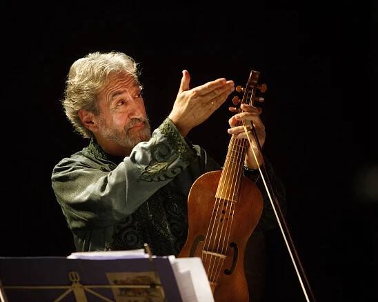 Jordi Savall, mit grauem Haar und einem grün gemusterten Hemd, hält ein hölzernes Saiteninstrument in der Hand. Er steht auf einer schwach beleuchteten Bühne. Im Vordergrund sind Notenblätter und ein Notenpult zu sehen.