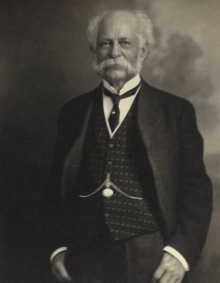 A formal, black-and-white portrait of an older man with a full white mustache and thick, wavy white hair. He is wearing a dark suit jacket, a patterned vest with a watch chain, a starched white collar, and a tie. He is looking directly at the camera with a neutral expression. The background is a simple, dark gradient.