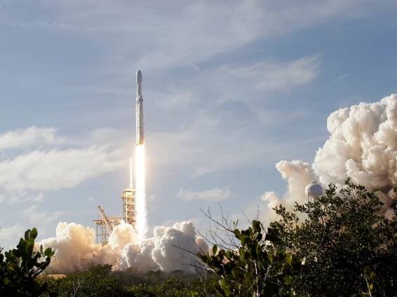 A rocket launches into a partially cloudy sky, forming large smoke billows underneath and to the right of the rocket.