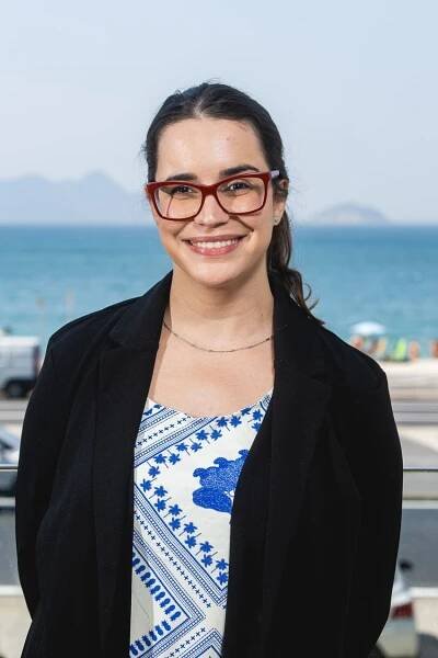 A photograph of Clarissa Carneiro shows her wearing a blue and white shirt with a black blazer. She has red, square-framed glasses and is smiling. Carneiro is the co-executive director at the Brazilian Reproducibility Network.