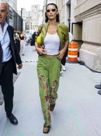 Bella Hadid wears a white tank top, green embroidered trousers, a green leather jacket, oval sunglasses, and a yellow shoulder bag