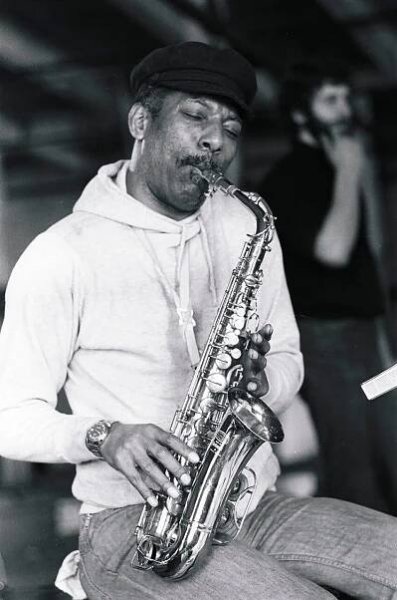A black-and-white photo of Gene Barge, a man wearing a white sweatshirt and a black cap, sitting and playing an alto saxophone.
